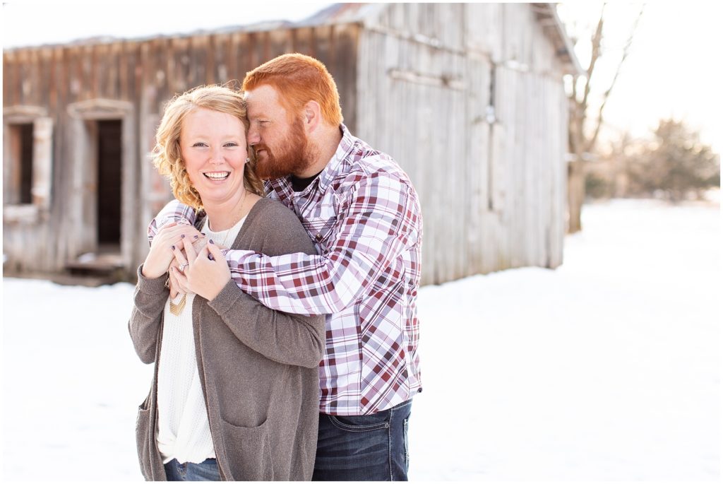 Engagement photos posed in snow near a rustic barn.